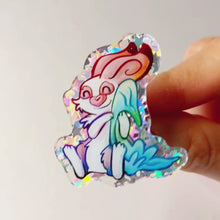 Load and play video in Gallery viewer, A pin gets tilted back and forth to better display the glittery acrylic. A glittery acrylic pin featuring a character from Rainbow of Crazy Comics: Linus an English spot rabbit, smiles as he cuddles a light blue blanket.
