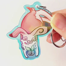 Load and play video in Gallery viewer, A clear, blue-tinted holographic acrylic keychain featuring a character from Rainbow of Crazy Comics: an English spot rabbit, lying down, admires a potted plant. 
