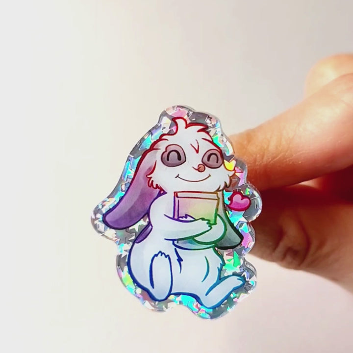 An acrylic pin featuring a character from Rainbow of Crazy Comics: a mini lop rabbit hugging a book, with a tiny heart next to him.