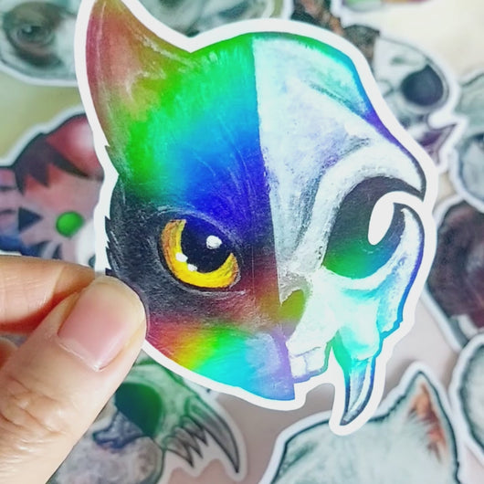 hand holding a cat/skull sticker and tilting it back and forth to show the holographic finish.