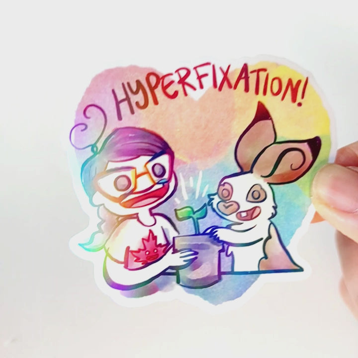 A vinyl rainbow holographic sticker featuring characters from Rainbow of Crazy Comics: Heart shaped sticker with the word "HYPERFIXATION!" above a Comic Jo and an English Spot rabbit, both smiling as they tend to a potted plant.