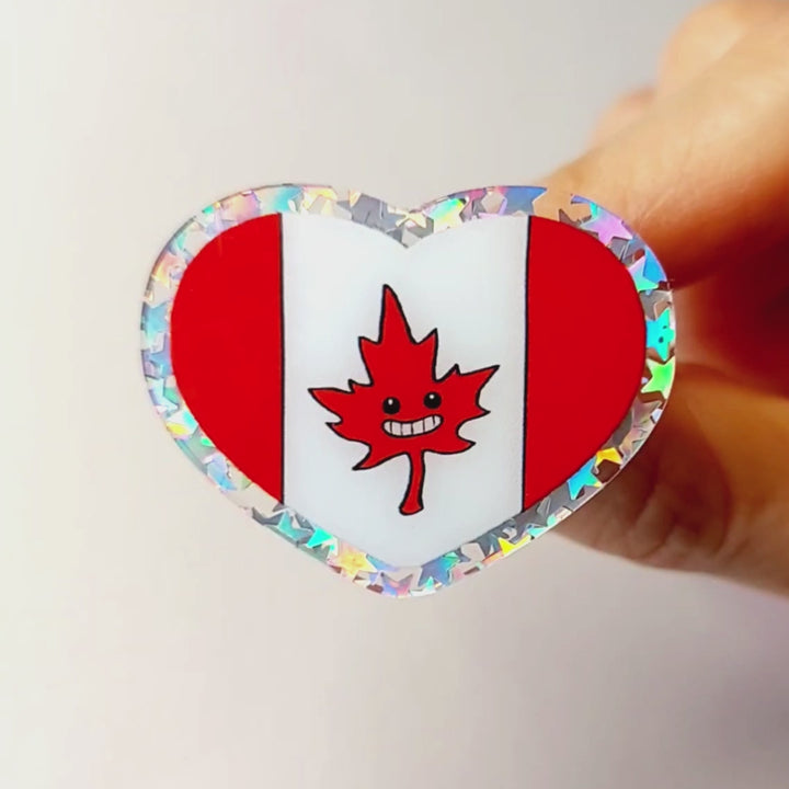 A glittery acrylic pin featuring a character from Rainbow of Crazy Comics: the Canada flag with a stylized maple leaf with happy face.