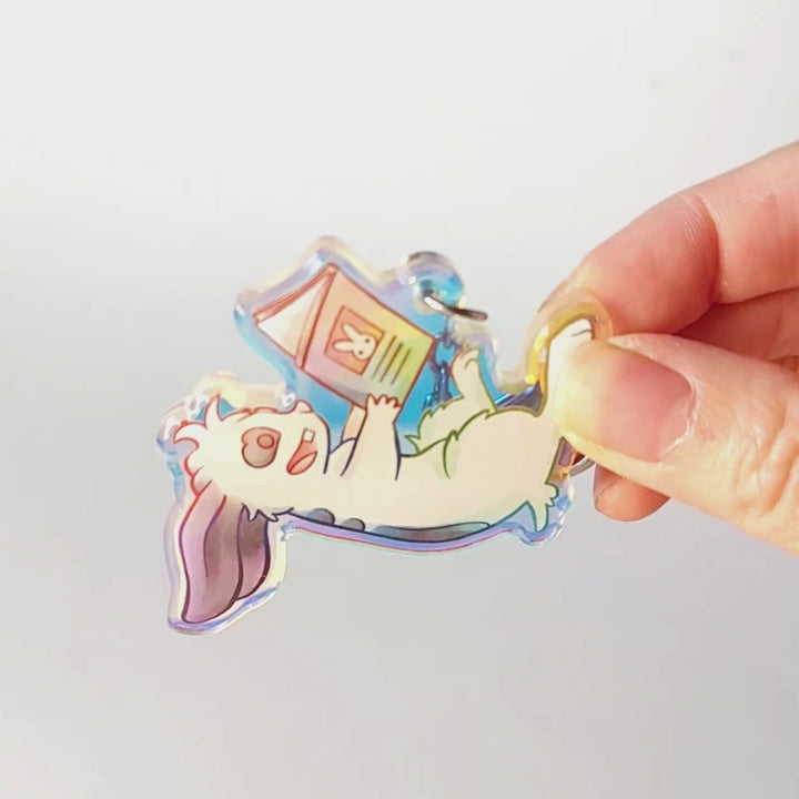A clear, rainbow holographic acrylic keychain featuring a character from Rainbow of Crazy Comics: a smiling mini lop rabbit lounges on his back as he reads a book with a picture of a rabbit on the back.