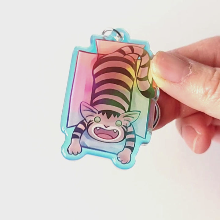 A clear, blue tinted holographic acrylic keychain featuring a character from Rainbow of Crazy Comics: a smiling tabby cat lounges in a cardboard box.