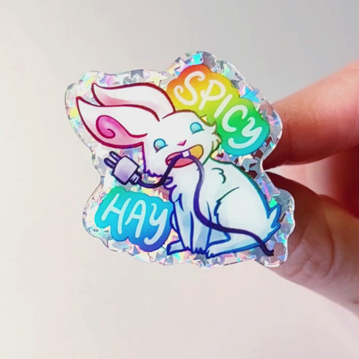 A glittery acrylic pin featuring a character from Rainbow of Crazy Comics: a smiling white rabbit holding a charging cable in her mouth, with the words "SPICY HAY in rainbow colours.