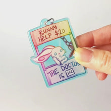Load and play video in Gallery viewer, A clear, rainbow holographic acrylic keychain featuring a character from Rainbow of a Crazy Comics: a smiling white rabbit at a homemade booth that says "bunny help $20" at the top, and "the doctor is in" at the bottom. 

