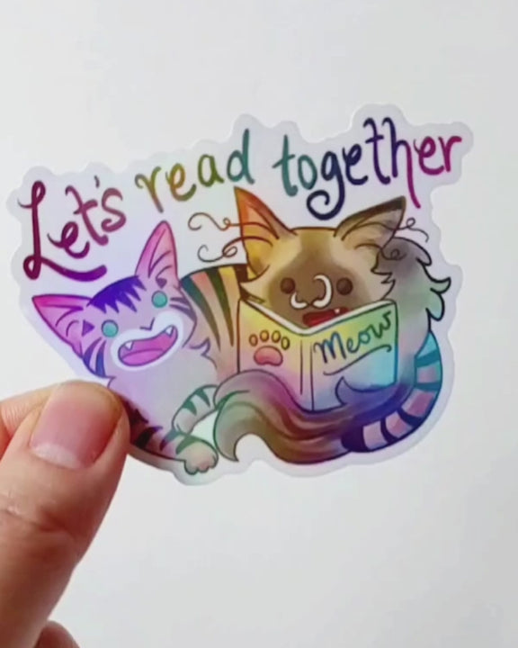 hand tilting a sticker to show its holographic finish. a sticker with two cats from Rainbow of Crazy Comics: a tabby cat next to a ragdoll cat that's holding a book titled "Meow". At the top of the sticker, it says, "Let's read together".