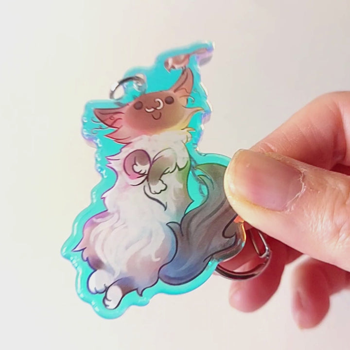 A clear, rainbow holographic acrylic keychain featuring a character from Rainbow of Crazy Comics: a smiling fluffy ragdoll cat, lying on her back, with a tiny black-capped chickadee, the bird perched on the cat's ear.