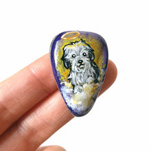 Load image into Gallery viewer, a beach stone hand painted with a sheepdog, as an angel with a halo, sitting on clouds against a purple and yellow sky. available as a keepsake or pendant necklace
