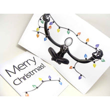 Load image into Gallery viewer, a greeting card with outside text: "Merry Christmas!" with a string of lights beneath / Inside features an illustration of Slender Man sitting cross-legged, arms out stretched, holding a long line of christmas lights

