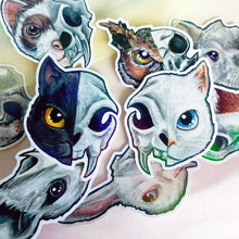 Load image into Gallery viewer, A collection of die-cut stickers featuring stylized animal skulls, including a cat/skull with blue eyes, a black cat/skull with yellow eyes, and other animal skull designs in the background.
