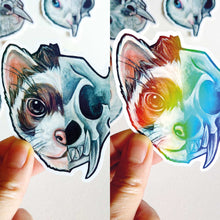 Load image into Gallery viewer, Split screen. A hand holds a ferret skull sticker. The sticker has normal colours on the left side. The right side has the sticker slightly tilted, showing its holographic finish.
