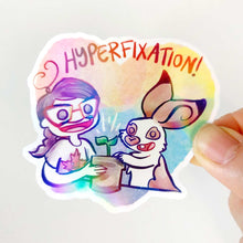 Load image into Gallery viewer, A vinyl rainbow holographic sticker featuring characters from Rainbow of Crazy Comics: Heart shaped sticker with the word "HYPERFIXATION!" above a Comic Jo and an English Spot rabbit, both smiling as they tend to a potted plant.
