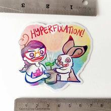 Load image into Gallery viewer, A vinyl rainbow holographic sticker featuring characters from Rainbow of Crazy Comics: Heart shaped sticker with the word "HYPERFIXATION!" above a Comic Jo and an English Spot rabbit, both smiling as they tend to a potted plant.
