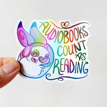 Load image into Gallery viewer, A vinyl rainbow holographic sticker featuring a character from Rainbow of Crazy Comics: a smiling rabbit wearing headphones. the sticker reads, "Audiobooks count as reading"
