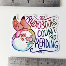 Load image into Gallery viewer, A vinyl rainbow holographic sticker featuring a character from Rainbow of Crazy Comics: a smiling rabbit wearing headphones. the sticker reads, "Audiobooks count as reading"
