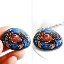 Load image into Gallery viewer, a small beach rock, hand painted with zodiac art: an orange and red crab, the sign of cancer, surrounded by blue water. available as a stone keepsake, magnet or pendant necklace
