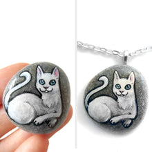 Load image into Gallery viewer, A small beach stone hand painted with a portrait of a white cat with blue eyes, available as a stone, magnet or pendant necklace.
