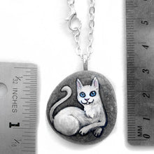 Load image into Gallery viewer, The white cat portrait necklace, hand painted on a small beach rock, next to try rulers to show its size: 1 1/4" x 1 1/8" or 3.1 cm x 2.8 cm 
