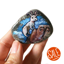 Load image into Gallery viewer, Rock art featuring a painting of a ram on a mountain, in front of a cloudy blue sky. The symbol for Aries is painted on the bottom. It's available as a keepsake, or as a fridge magnet.
