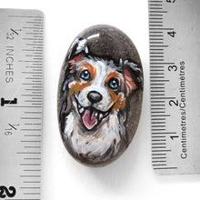 Load image into Gallery viewer, A small rock, hand painted with art of a smiling Australian Shepherd, next to twonrulers to show its size: 1 9/16" x 15/16" or 4 cm x 2.4 cm

