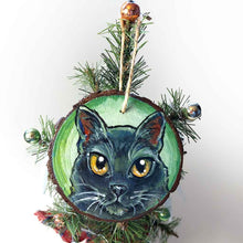 Load image into Gallery viewer, a round wood ornament, hand painted with a portrrait of a black cat's face, with yellow eyes, against a soft green background
