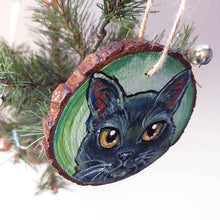 Load image into Gallery viewer, a round wood ornament, hand painted with a portrrait of a black cat's face, with yellow eyes, against a soft green background
