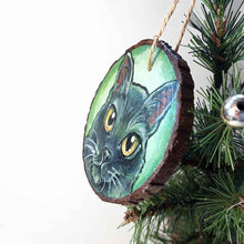 Load image into Gallery viewer, a round wood ornament, hand painted with a portrrait of a black cat's face, with yellow eyes, against a soft green background
