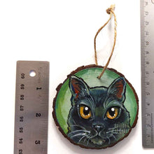Load image into Gallery viewer, a round wood ornament, hand painted with a portrrait of a black cat's face, with yellow eyes, against a soft green background
