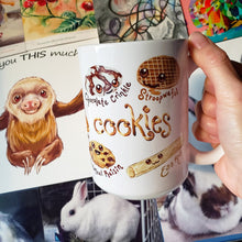 Load image into Gallery viewer, A hand holding up the 15 oz Cookie Lovers Smile Mug, in front of a wall with various art and photos.
