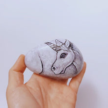 Load and play video in Gallery viewer, a stone hand painted with the portrait of a unicorn with purple eyes, its legs on the underside, and tail on the back
