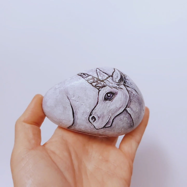 a stone hand painted with the portrait of a unicorn with purple eyes, its legs on the underside, and tail on the back