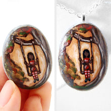 Load image into Gallery viewer, small rock art: a beach stone is hand painted with a girl in red, sitting on a tree swing, available as either a keepsake, fridge magnet, or a necklace
