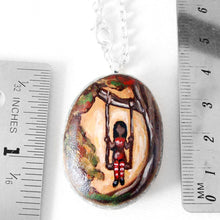 Load image into Gallery viewer, a beach rock with art of a woman in red, sitting on a tree swing. available as a necklace, the stone is next to two rulers to show its size: 1 1/4" x 1 1/8" or 3.1 cm x 2.8 cm
