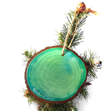 Load image into Gallery viewer, a christmas wood ornament, hand painted with the portrait of a syrian hamster, standing on the snow, watching snowflakes fall against a green and yellow sky, signed on the back with, Rainbow of Crazy
