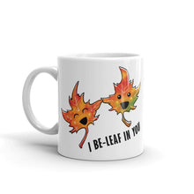 Load image into Gallery viewer, A 11 oz white ceramic mug, printed with art of two maple leaves with happy faces, and the words. "I be-leaf in you"
