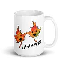Load image into Gallery viewer, A 15 oz white ceramic mug, printed with art of two maple leaves with happy faces, and the words. "I be-leaf in you"
