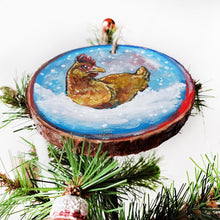 Load image into Gallery viewer, A holiday ornament with art of a chicken, sitting on snow as snowflakes fall. The ornament is painted in blues and reds.
