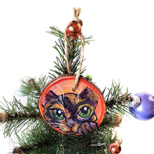 Load image into Gallery viewer, A wood Christmas tree ornament, hand painted with art of a tortoiseshell cat, with green eyes, in front of a red and pink background

