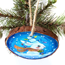 Load image into Gallery viewer, A wood Christmas ornament, hand painted with a white bunny rabbit sitting on the snow with a blue background, with an orange scarf, green mittens, and christmas lights on its ears
