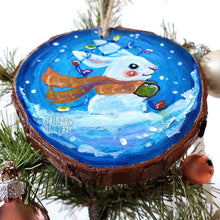 Load image into Gallery viewer, A wood Christmas ornament, hand painted with a white rabbit sitting on the snow with a blue background, with an orange scarf, green mittens, and christmas lights on its ears
