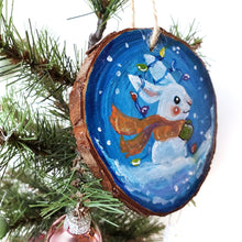 Load image into Gallery viewer, A wood Christmas ornament, hand painted with a white rabbit sitting on the snow with a blue background, with an orange scarf, green mittens, and christmas lights on its ears
