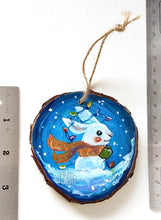 Load image into Gallery viewer, A wood Christmas ornament, hand painted with a white bunny rabbit sitting on the snow with a blue background, with an orange scarf, green mittens, and christmas lights on its ears
